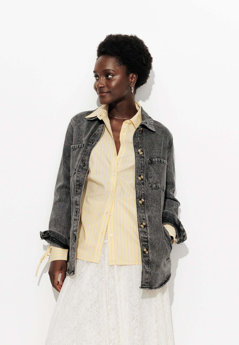 Veste en denim foncé avec un design à col, fermeture à boutons et poche poitrine, portée sur une chemise à rayures jaunes claires à boutons et une jupe en dentelle blanche.