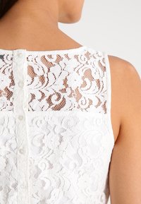 Haut en dentelle blanche avec un motif floral, doté d'une fermeture à boutons à l'arrière et d'une texture douce, créant un design superposé.