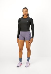 Kvinde står iført en sort langærmet top, grå træningsshorts, blå sokker og lavendel løbesko på en hvid baggrund.