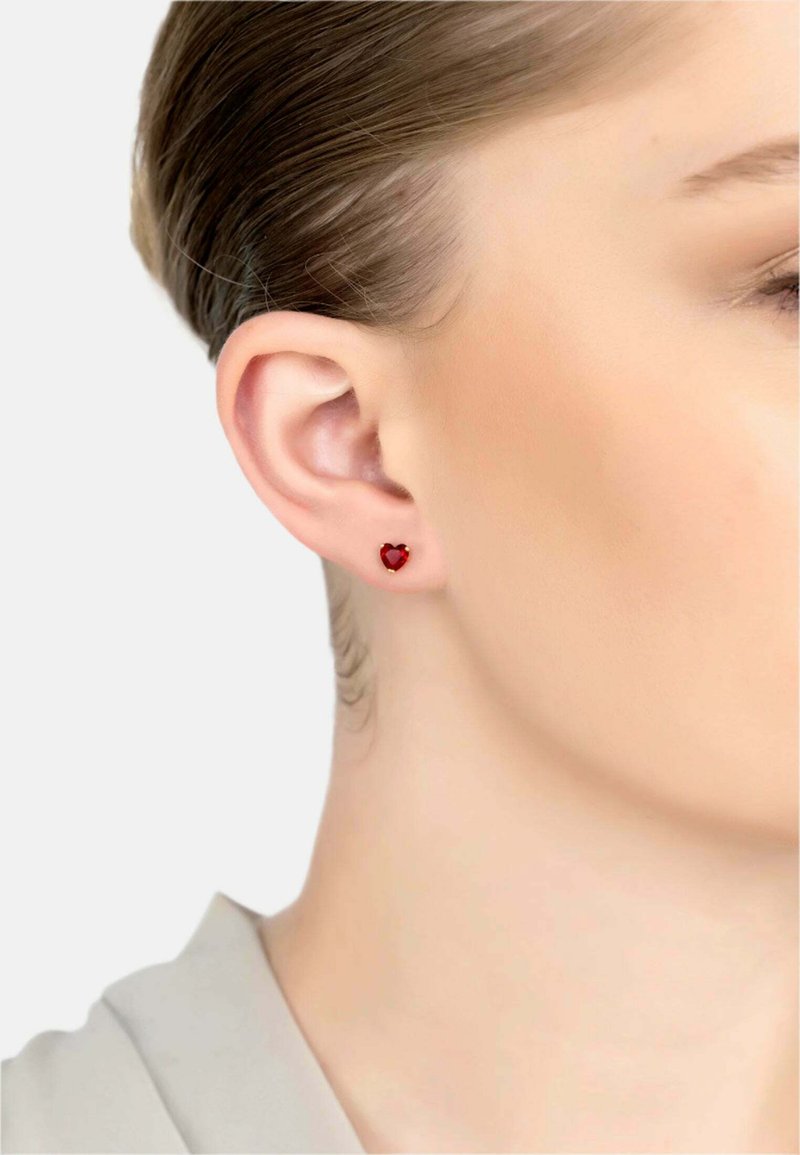 Rote herzförmige Ohrstecker aus glänzendem Material, vor einem hellen Hautton und einer schlichten beigen Kleidung.