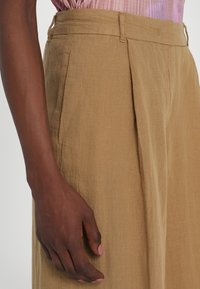 Linen trousers in a warm brown color, featuring two front pleats, a classic waistband, and detailed stitching on the side pockets.