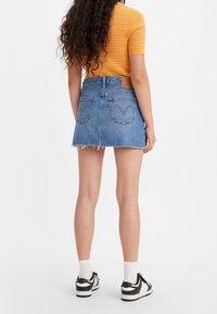 Denimminikjol med rådkant, blå färg, två bakfickor och en läderlogotyp vid midjan. Modell bär svarta sneakers.