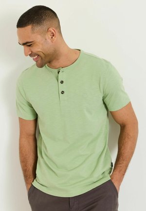 Smiling man with short hair wearing light green short-sleeve Henley shirt and dark pants, hands in pockets, looking down to left.