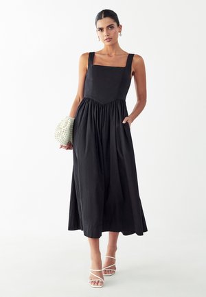 Mujer vistiendo un vestido midi negro sin mangas con escote cuadrado, sosteniendo un bolso de mano blanco con cuentas y llevando sandalias blancas de tiras.