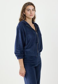 Marineblauwe velours hoodie met rits, losse pasvorm, verstelbare capuchon met trekkoord en geribbelde boorden. Zachte textuur en gouden ritsdetail.