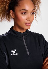 Schwarzes, zip-up Sportoberteil mit hohem Kragen, strukturiertem Stoff und grauen Schulterakzenten, die das Hummel-Logo in Weiß aufweisen.