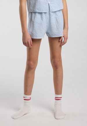 Person wearing light blue and white striped shorts and matching top, with white socks featuring two red stripes, standing on a plain background.