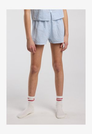 Personne portant un short rayé bleu clair et blanc avec un haut assorti, avec des chaussettes blanches à deux bandes rouges, debout sur un fond uni.