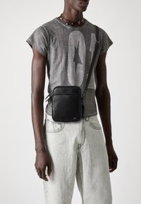 Borsa a tracolla in pelle nera con due scomparti zip, forma rettangolare e tracolla regolabile. Indossata con una maglietta grafica grigia e jeans chiari.