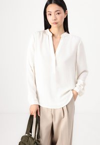 Hvit langermet bluse med en liten krage og en knappestolpe. Kombinert med beige bukser, holder modellen en grønn håndveske. Glatt tekstur.
