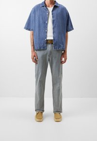 Persona vestida con una camisa de mezclilla azul de manga corta sobre una blusa blanca, pantalones a rayas, cinturón marrón y zapatos de lona beige, de pie contra un fondo liso.