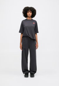 Schwarzes Baumwoll-T-Shirt mit kleinem rotem Logo, kombiniert mit locker sitzenden schwarzen Jogginghosen. Modell steht selbstbewusst in schwarzen Sneakers.