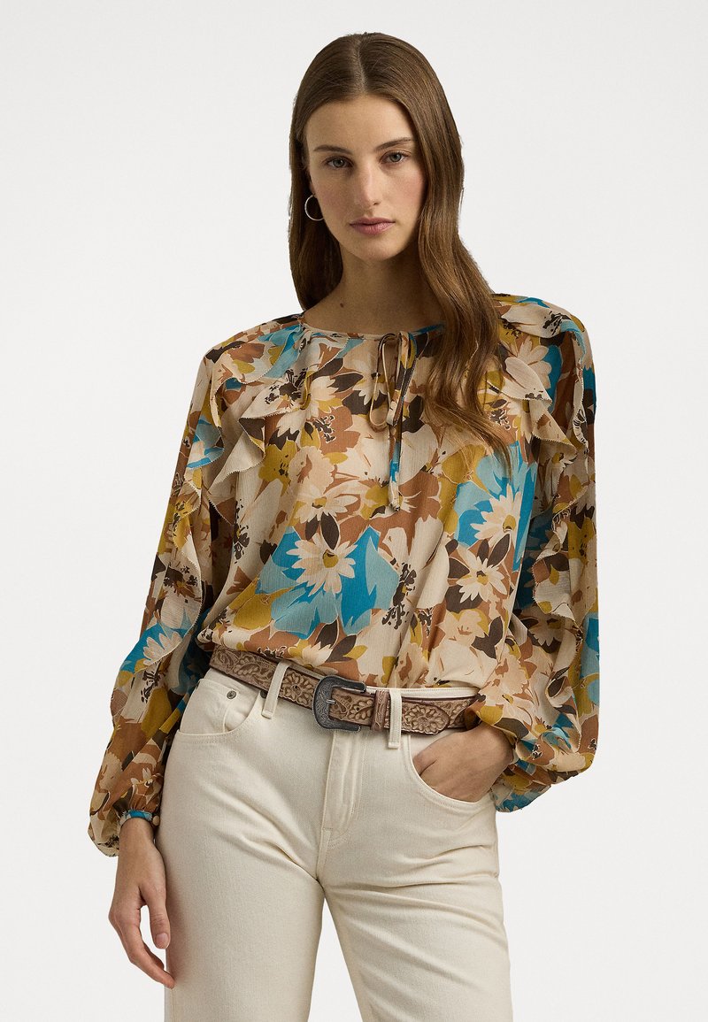 Blomstrete bluse med båt hals, volanger på skuldrene og lange ermer i beige, blå, gul og brun. Kombinert med hvite jeans.