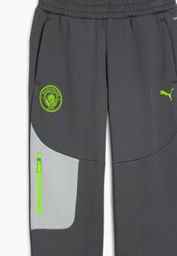 Szare dresy wykonane z teksturowanej tkaniny, z neonowo-zielonym logo Manchester City oraz odblaskowym kieszeniem na suwak z boku.