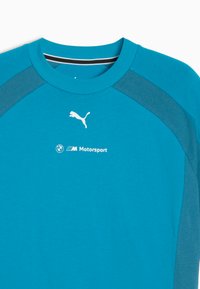Turkos träningsskjorta med kontrasterande mörka ärmar. Har vit Puma-logotyp och BMW Motorsport-märkning på framsidan. Slät struktur.