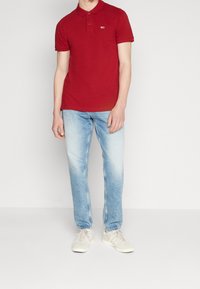 Červená bavlněná polo košile s dvěma knoflíky a vyšitým logem, doplněná světle modrými rovnými džíny a b ílými teniskami.