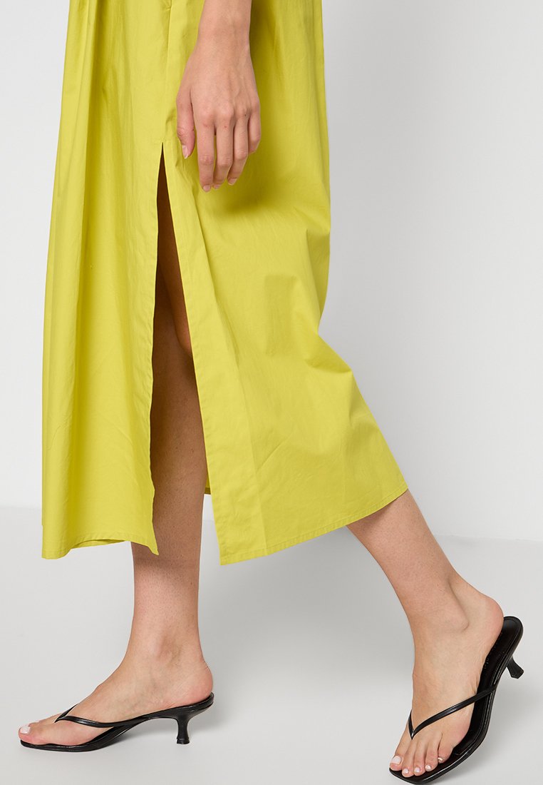 Gul, knälång klänning med rak skärning och sidoklyftor, kombinerad med svarta, klacksandaler. Slät, lätt tygstruktur.