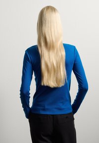 Long-sleeve blue top made of smooth fabric, paired with dark trousers. The back view shows a sleek design and straight, long blonde hair.