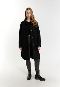 Černý vlněný kabát s knoflíky, velkými kapsami a oversize střihem, doplněný o tmavé džíny a černé kotníkové boty. Hladká textilie.