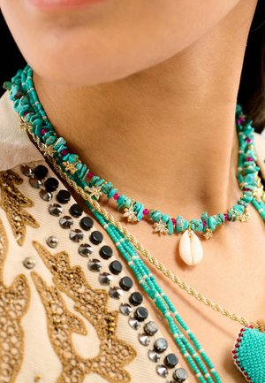 Collier de perles avec des pierres turquoise, des accents roses et des détails dorés en forme d'étoile, orné d'un pendentif en coquillage. Superposé sur un tissu à motifs.