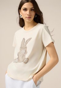 T-shirt en coton crème avec des manches courtes, orné d'un design de lapin scintillant en strass sur le devant. Texture lisse, coupe décontractée.