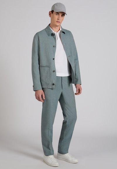 Mężczyzna w jasnoszarym garniturze, białej koszulce polo, szarej czapce i białych trampkach, stojący na jednolitym tle.