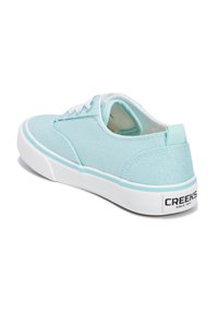 Zapatilla de lona azul clara con suela de goma blanca, diseño de cordones y marca en el talón, mostrando una parte superior texturizada y acentos en contraste.