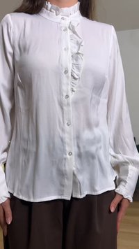 Bela bluza z dolgimi rokavi z nabranim ovratnikom in sprednjo vrstico z gumbi, kombinirana s temnordečimi hlačami, ki jo nosi stoječa oseba.