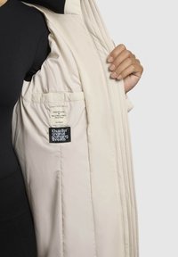 Lyse beige pufferjakke med en glat, quiltet tekstur. Interiørlabels synlige, herunder "Made with Love" og "Khujo."