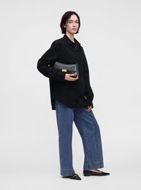 Personne portant une chemise noire à motifs, un jean bleu large, et des ballerines noires pointues, tenant une pochette noire sur un fond uni.