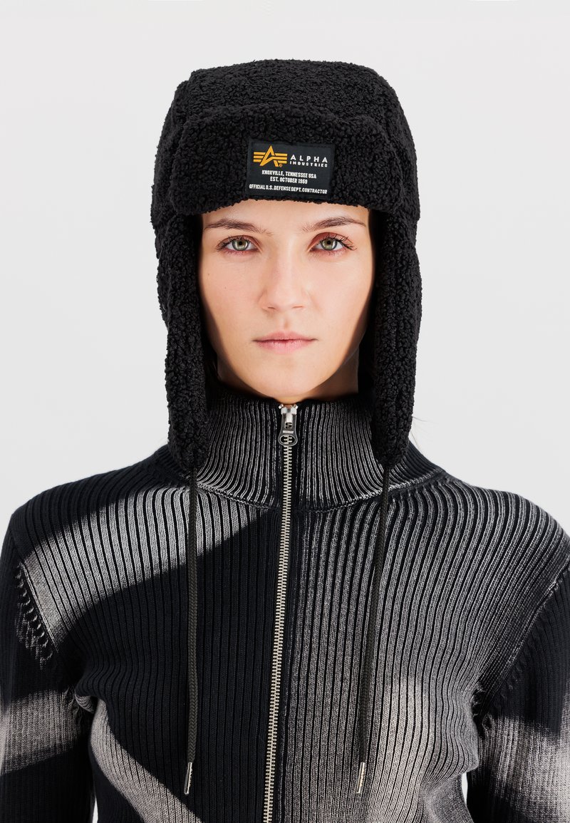 Vrouw die naar voren kijkt draagt een zwarte fleece trappersmuts met een voorlabel en een zwart-grijze geribbelde trui met rits.