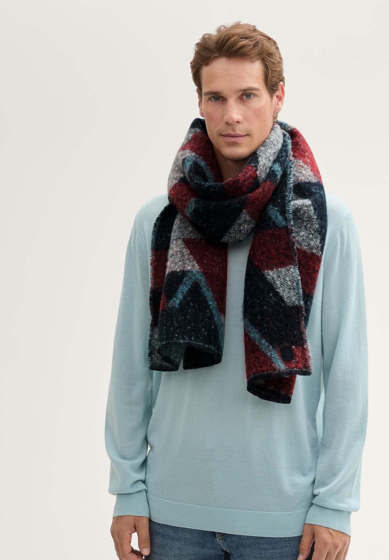 Écharpe multicolore avec un motif géométrique en rouge, marine, gris et turquoise. Fabriquée en tissu doux avec un bord effiloché et un détail d’étiquette.