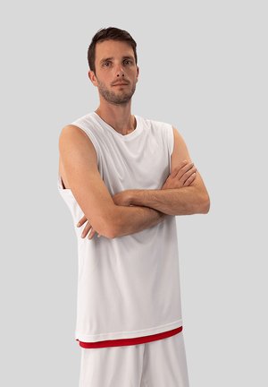 Mann mit kurzen braunen Haaren, der ein weißes ärmelloses Sportshirt und weiße Shorts trägt, steht mit verschränkten Armen vor grauem Hintergrund.