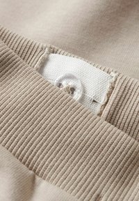 Tejido texturizado beige con un patrón acanalado, que presenta una etiqueta de tela blanca con un botón claro. Vista en primer plano del detalle de la costura.