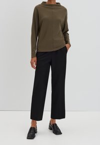 Haut long à col montant de couleur vert olive et texture, associé à un pantalon ample noir et des mocassins à talons noirs. Fond uni.