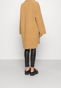 Cappotto beige testurizzato con una vestibilità rilassata, colletto oversize e spalle cadenti, indossato sopra leggings in pelle nera e scarpe con plateau.
