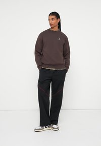 Joven con cabello trenzado, vestido con una sudadera marrón, pantalones de chándal negros con acentos rojos y zapatillas blancas, de pie contra un fondo blanco.