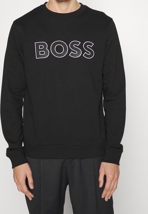 Man draagt een zwarte sweatshirts met "BOSS" in grote witte letters met omlijning op de borst, gecombineerd met donkere broek.