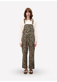 Vrouw staat rechtop en kijkt naar voren, gekleed in een beige langemouwen top, een overall met luipaardprint en bruine gesloten schoenen op een lichte achtergrond.