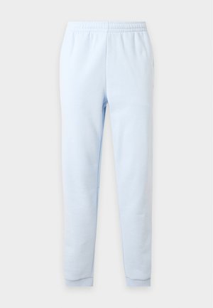Homme portant un t-shirt gris Lacoste avec un logo crocodile vert, un pantalon de jogging bleu clair et des baskets blanches, debout les mains dans les poches.