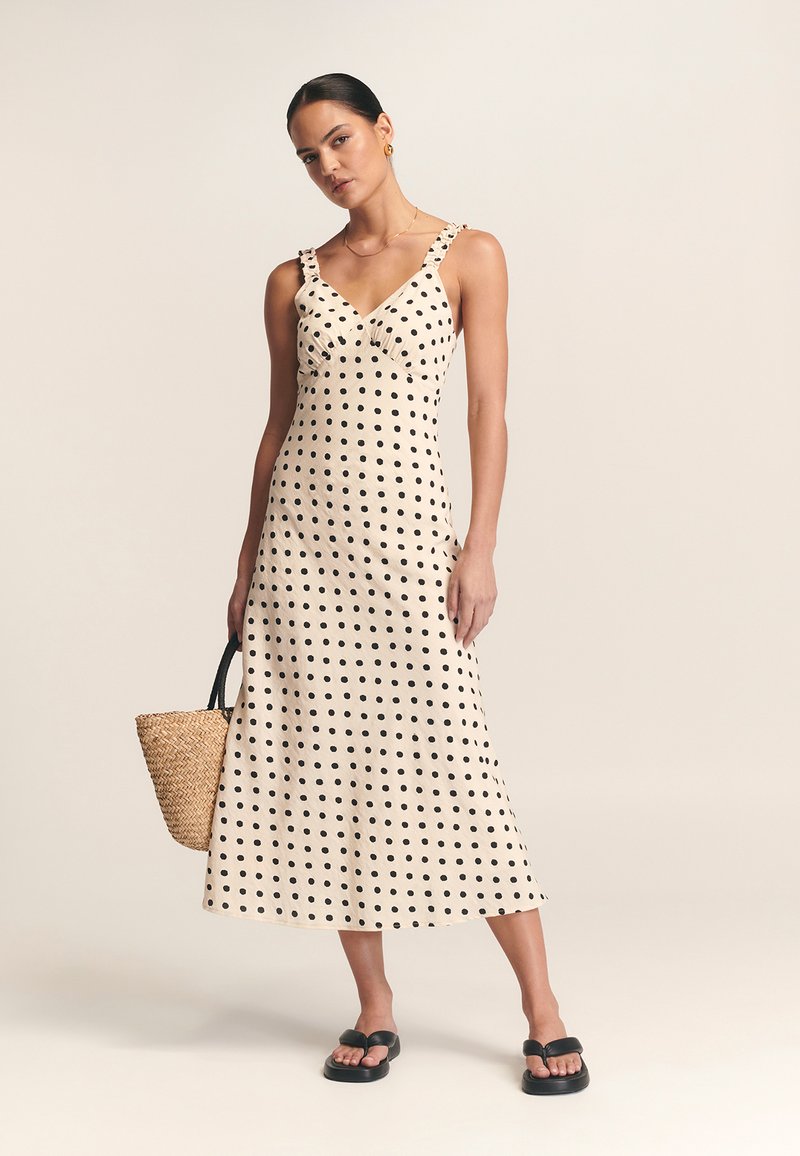 Romantische midi-jurk met polka dot in crème met zwarte stippen, voorzien van een v-hals, gerimpelde schouderbanden en een vloeiende silhouet. Gemaakt met een geweven tas en platte sandalen.
