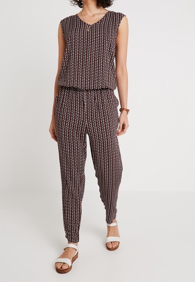 Combinaison sans manches à motifs avec un haut ample et un pantalon fuselé, portée avec des sandales blanches et un collier simple sur une personne debout.