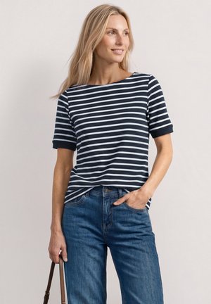 Kvinde med langt blondt hår bærer en marineblå og hvid stribet t-shirt med korte ærmer og blå jeans, og holder en brun håndtaske i den ene hånd.