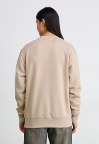 Felpa beige con colletto a giro e maniche lunghe, realizzata in materiale morbido con polsini e orlo a coste, indossata con jeans sbiaditi.
