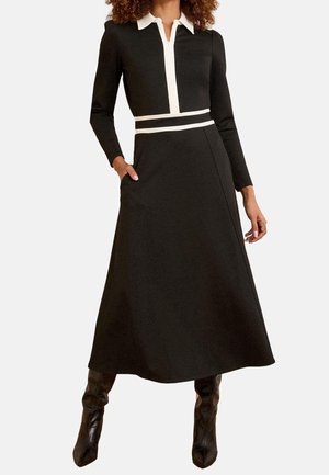 Femme portant une longue robe noire avec un col blanc et des bandes à la taille, posant avec une main dans la poche et des bottes noires montantes jusqu'au genou.
