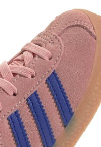 Gros plan sur une basket en cuir suédé rose avec des lacets roses, trois bandes zigzag bleues sur le côté et une semelle en caoutchouc translucide.