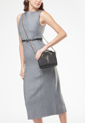 Donna in vestito midi grigio senza maniche con cintura nera, che tiene una borsa nera trapuntata con tracolla a catena d'oro e stemma dorato.