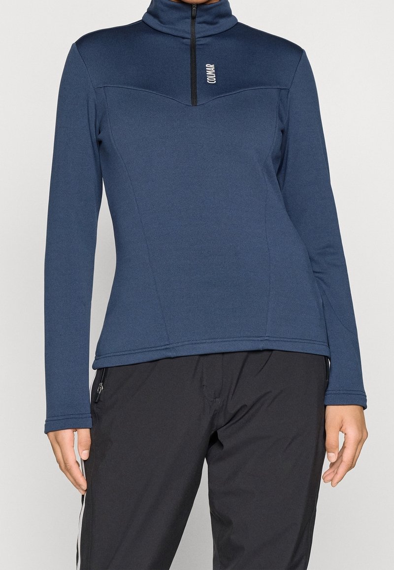 Femme portant un haut zippé bleu marine à manches longues et un pantalon noir avec des fermetures éclair latérales sur un fond uni.