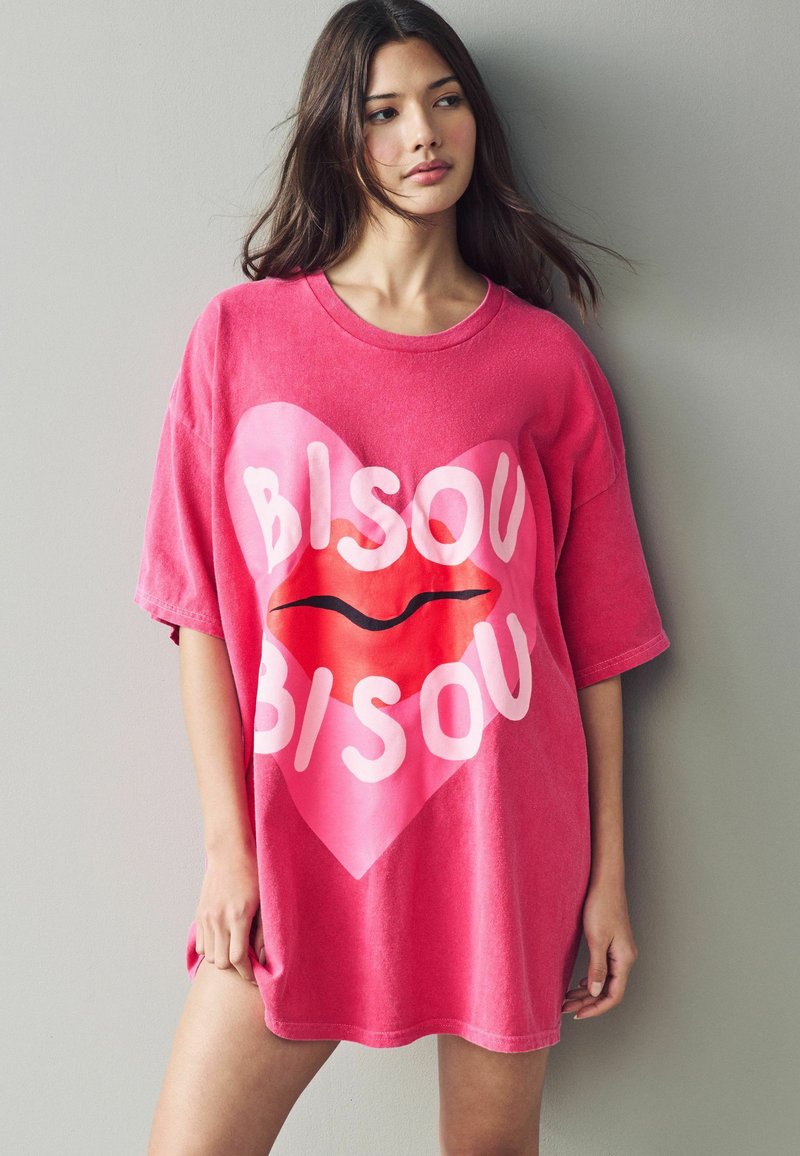 T-shirt oversize rosa con un cuore rosa chiaro e grafica di labbra rosse con la parola "BISOU" in lettere bianche in grassetto. Texture morbida.