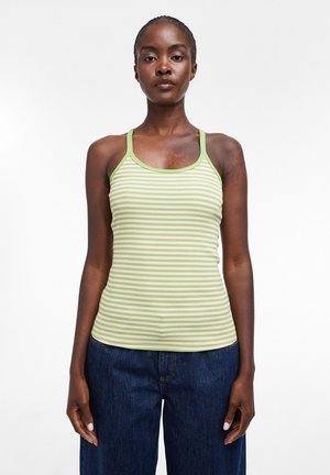 Femme à la peau foncée portant un haut sans manches rayé vert et blanc et un jean bleu, debout devant un fond blanc uni.